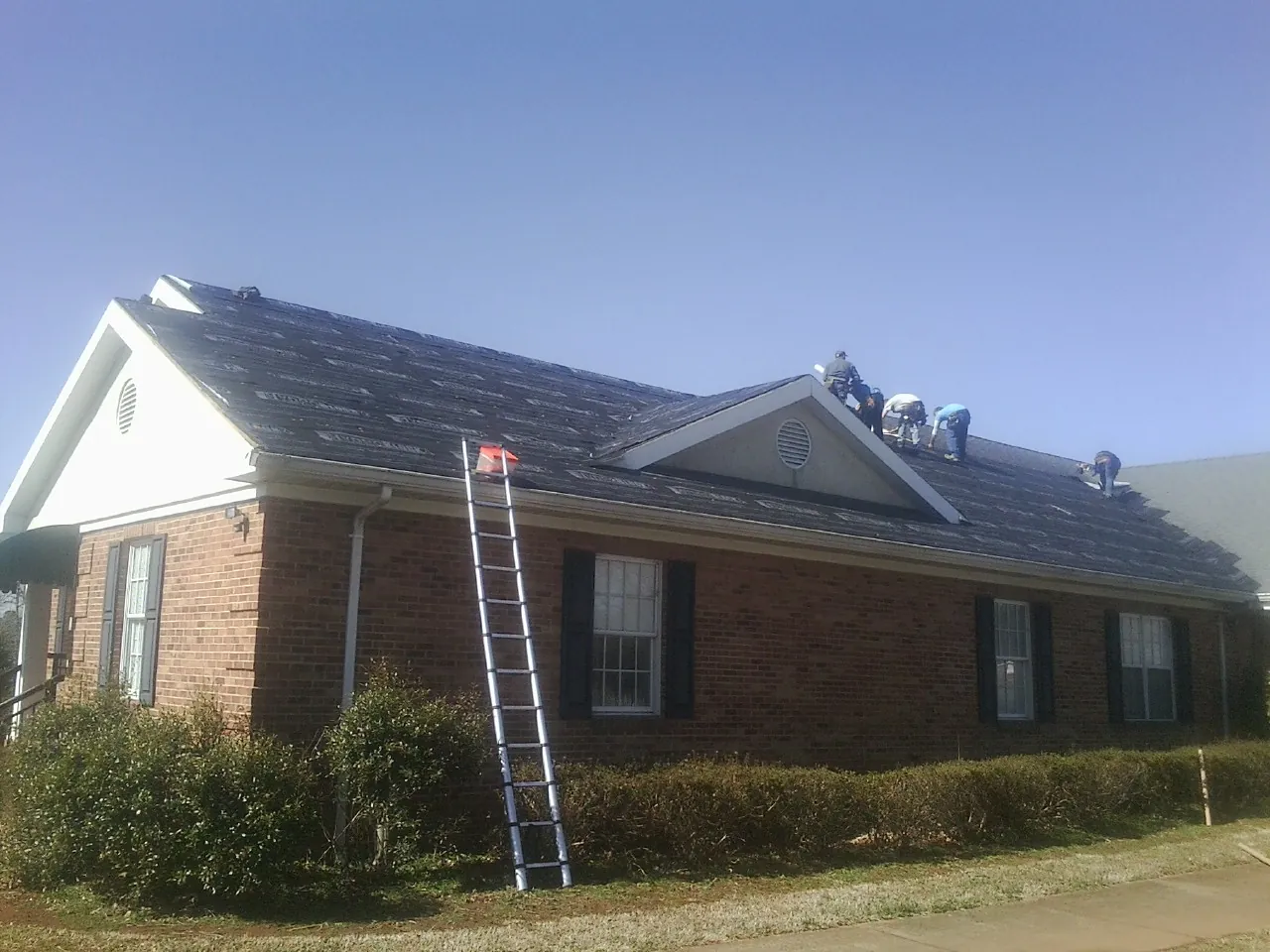 Crew tearing shingles off a brick ranch roof with a ladder set — Emergency Roof Repair work by our crew in Glen Flora