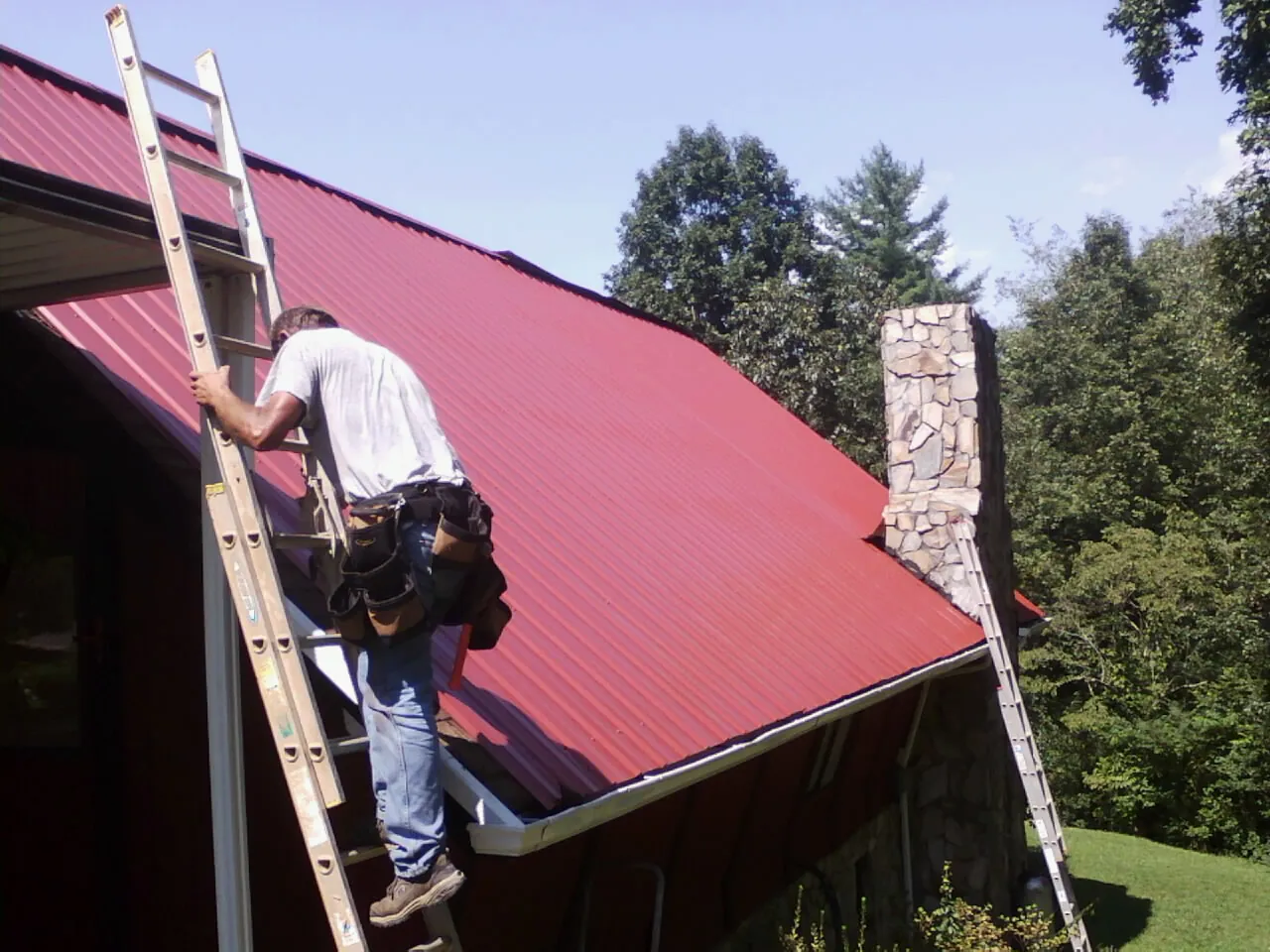 Roofer on a ladder beside a red metal roof and stone chimney — Soffit & Fascia Repair by Roofing Brothers in Glen Flora